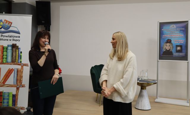 Na zdjęciu znajdują się dwie kobiety, jedna z mikrofonem. Kobiety stoją przed sceną, na której znajduje się fotel, plakat promujący spotkanie, oraz baner po lewej stronie z książkami i logiem biblioteki.