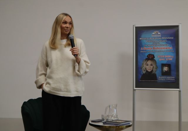 Na zdjęciu znajduje się kobieta stojąca na scenie z mikrofonem, obok niej stoi plakat promujący spotkanie.