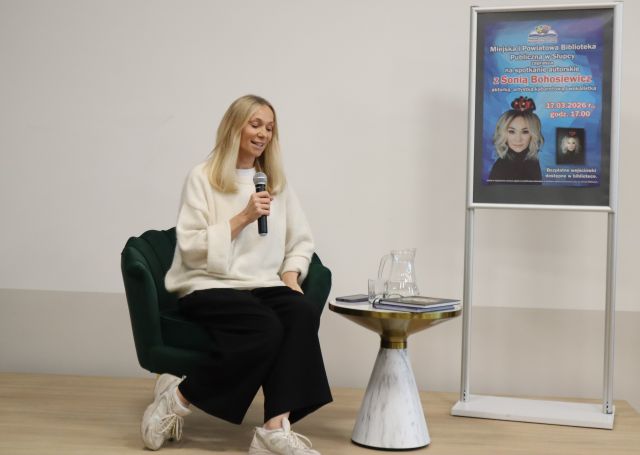 Na zdjęciu znajduje się siedząca na scenie kobieta z mikrofonem, obok niej stoi mały stolik z wodą oraz plakat promujący spotkanie.