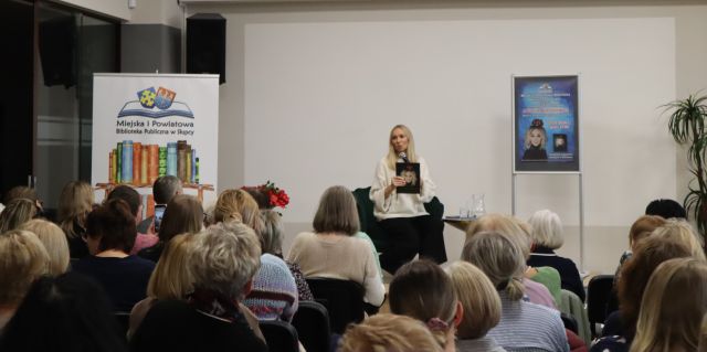 Na zdjęciu znajdują się ludzie na widowni skierowani w stronę kobiety na scenie, która trzyma w ręce swoją książkę. Na scenie znajduje się również plakat promujący spotkanie, a przy scenie po lewej stronie stoi baner z książkami i logiem biblioteki.