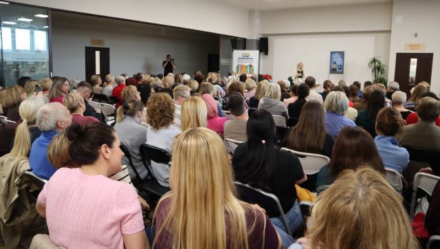 Na zdjęciu znajduje się widownia skierowana w stronę sceny, na której siedzi kobieta. Spotkanie odbywa się na holu biblioteki. 