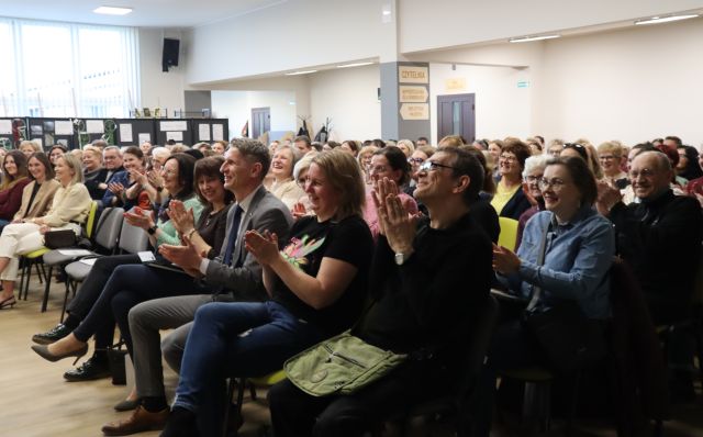 Na zdjęciu znajduje się około sto osiemdziesiąt osób siedząca na krzesłach na holu biblioteki i klaskają.