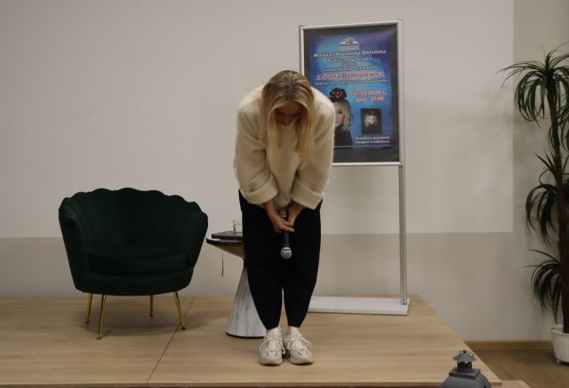 Na zdjęciu znajduje się kobieta na scenie, która się kłania, za nią stoi fotel, mały stolik oraz plakat promujący spotkanie.