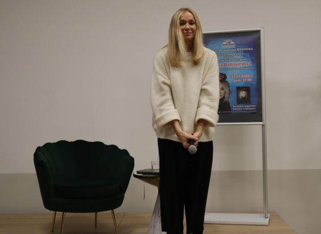 Na zdjęciu znajduje się kobieta stojąca na scenie, ma przed sobą mikrofon. Za kobietą na scenie stoi fotel oraz plakat promujący spotkanie.