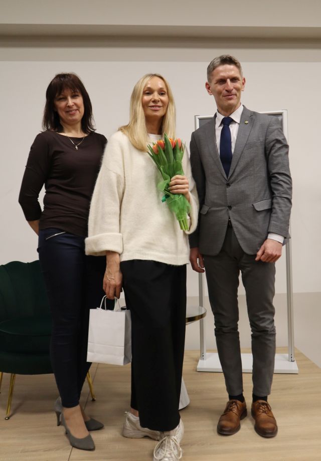 Na zdjęciu znajdują się dwie kobiety oraz jeden mężczyzna, kobieta na środku ma kwiaty i wspólnie stoją do zdjęcia. W tle znajduje się fotel oraz plakat promujący spotkanie.