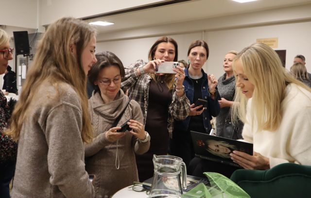 Na zdjęciu znajduje się kobieta podpisująca książkę oraz ludzie dookoła niej, jedna ze stojących kobiet robi zdjęcie.
