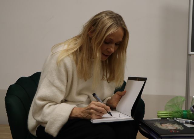 Na zdjęciu znajduje się siedząca na fotelu kobieta i podpisująca książkę.