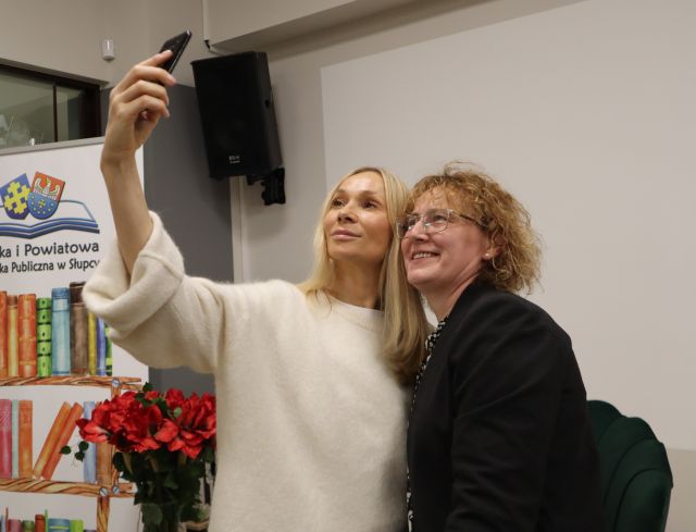 Na zdjęciu znajdują się dwie kobiety, jedna z nich wyciąga rękę aby zrobić zdjęcie. W tle po lewej znajduje się baner z książkami i logiem biblioteki.