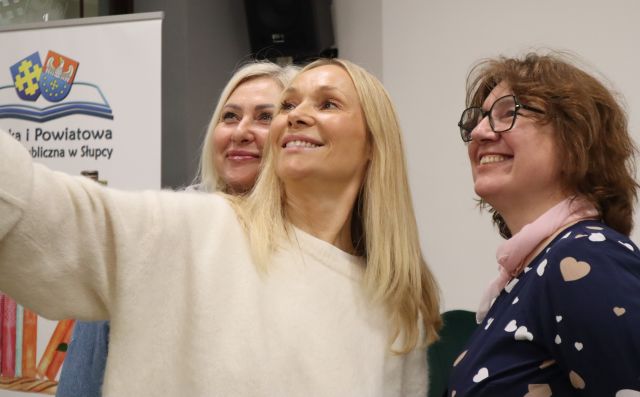 Na zdjęciu znajdują się trzy kobiety, kobieta po środku wyciąga rękę z telefonem i robi wspólne zdjęcie. Po lewej stronie za kobietami znajduje się baner z książkami i logiem biblioteki.