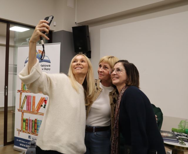 Na zdjęciu znajdują się trzy kobiety, jedna z nich ma wyciągniętą rękę i robi im wspólne zdjęcie, w tle po lewej znajduje się baner z książkami i logiem biblioteki.