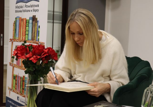 Na zdjęciu znajduje się kobieta siedząca na fotelu i wpisująca się do kroniki, za nią po lewej stronie znajdują się kwiaty i baner z książkami.