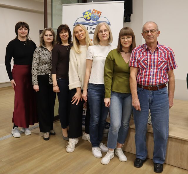 Na zdjęciu znajduje się sześć kobiet oraz jeden mężczyzna i stoją do wspólnego zdjęcia na tle baneru z książkami i logiem biblioteki.