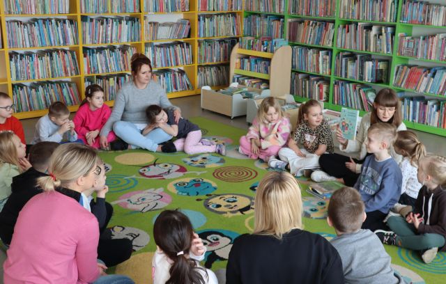 Na zdjęciu znajdują się przedszkolaki siedzące na dywanie w okręgu wraz z czterema kobietami, jedna z nich pokazuje trzymaną przez siebie książkę. W tle znajdują się półki z książkami.