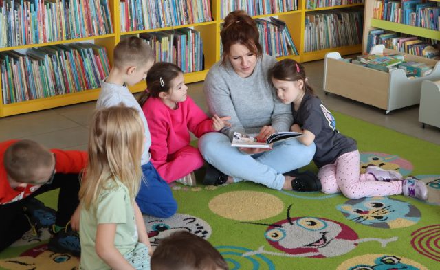 Na zdjęciu znajduje się piątka przedszkolaków na dywanie wraz z kobietą, która czyta im fragment z książki. W tle znajdują się półki z książkami.