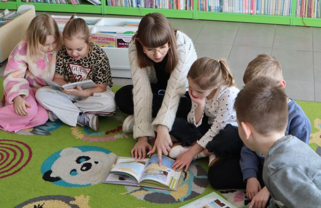Na zdjęciu znajduje się piątka przedszkolaków na dywanie, dwie dziewczynki przeglądają wspólnie jedną książkę, a trójka wraz z kobietą oglądają inną książkę, która leży na dywanie. W tle znajdują się małe półki z książkami.