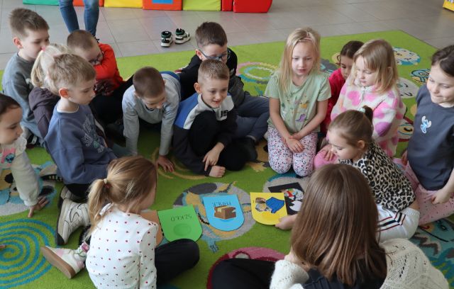 Na zdjęciu znajduje się grupa przedszkolaków na dywanie wraz z kobietą, na środku znajdują się wycięte z papieru kolorowe kosze.