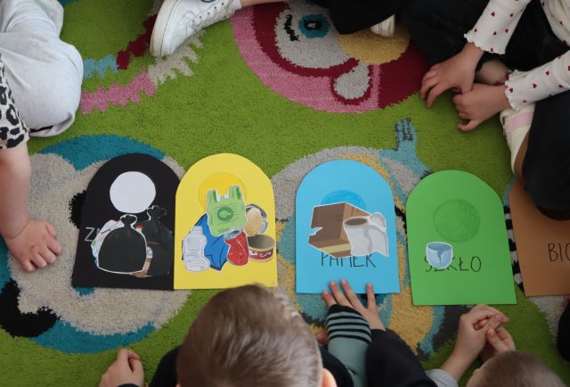 Na zdjęciu znajdują się wycięte z papieru kolorowe kosze, na których leżą Ilustracje śmieci, dookoła nich na dywanie siedzą pzedszkolaki.