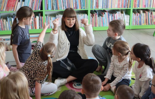 Na zdjęciu znajdują się przedszkolaki na dywanie, a na środku siedzi kobieta i pokazuje im dwie małe ilustracje. W tle znajdują się półki z książkami.