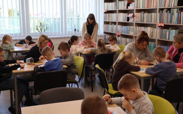 Na zdjęciu znajdują się przedszkolaki przy pięciu stolikach, przy których kolorują, na końcu przy jednym stoliku stoi kobieta. W tle znajdują się okna, a po prawej stronie półki z książkami.