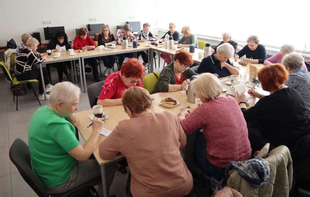 Na zdjęciu znajduje się dwadzieścia kobiet, które siedzą przy załączonych w literę C stolikach i rozwiązują zadania przy kawie. Warsztaty odbywają się w sali komputerowej.