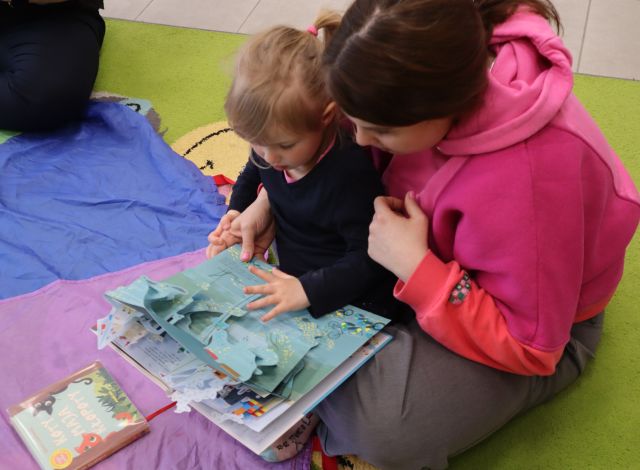 Na zdjęciu znajduje się kobieta z dzieckiem i przeglądają wspólnie książkę z wystającymi elementami. Przed nimi znajduje się kolorowa chusta.