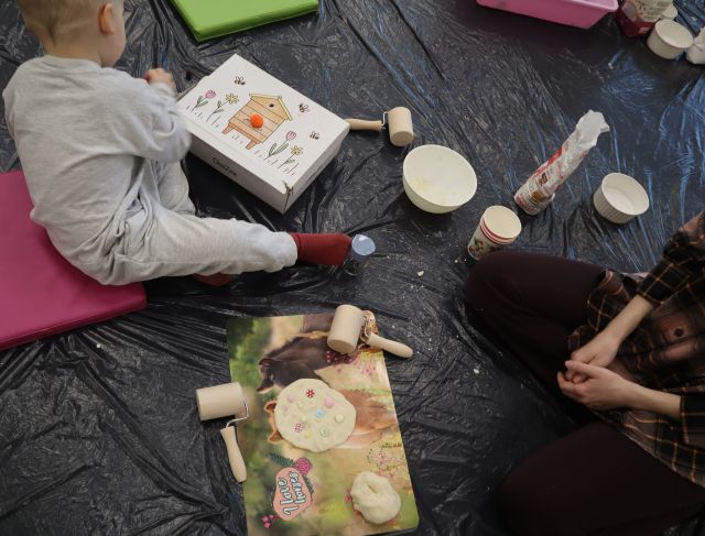 Na zdjęciu znajdują się masa solna, kubeczki, miska oraz chłopiec przed którym leży pudełko, a po prawej stronie znajduje się fragment siedzącej kobiety.