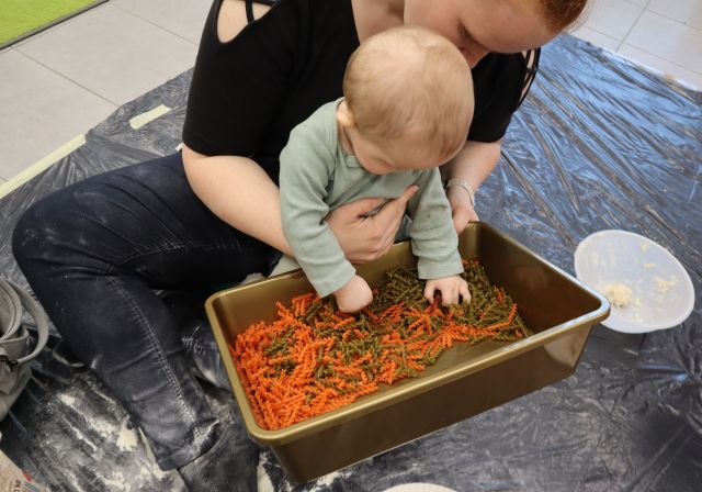 Na zdjęciu znajduję się kobieta z niemowlakiem, który bawi się kolorowym makaronem, który znajduje się w prostokątnym pojemniku. Na podłodze rozłożona jest ciemna folia.