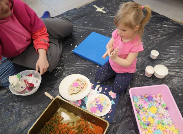 Na zdjęciu znajduje się dziewczynka bawiąc się masą solną, dookoła niej leżą talerzyki, pojemnik z ozdobami, a po lewej stronie siedzi kobieta.