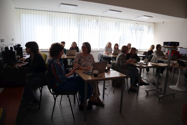 Na zdjęciu znajdują się kobiety siedzące przy stolikach z laptopami, otaz przy komputerach. Na środku znajduje się rzutnik. Warsztaty odbywają się w sali komputerowej, w tle znajdują się okna.