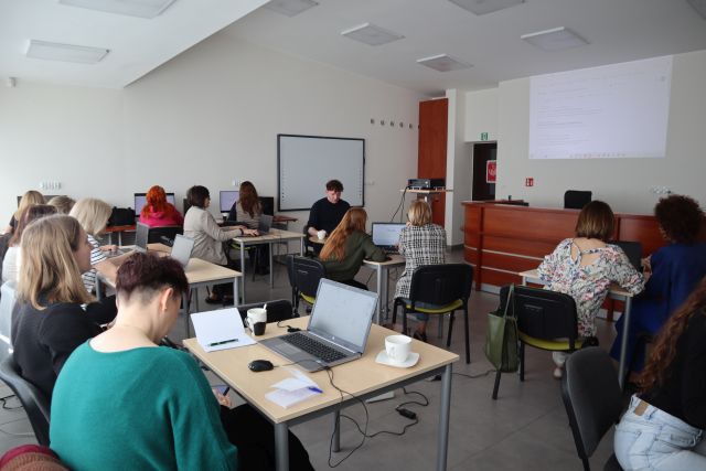 Na zdjęciu znajdują się kobiety oraz jeden mężczyzna przy stolikach z laptopami oraz przy trzech komputerach. Po prawej stronie na ścianie wyświetlana jest prezentacja o sztucznej inteligencji. Warsztaty odbywają się w sali komputerowej.