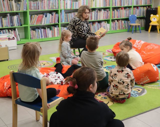 Na zdjęciu znajdują się dzieci na dywanie, pufach a przed nimi siedzi kobieta z książką. W tle znajdują się półki z książkami.