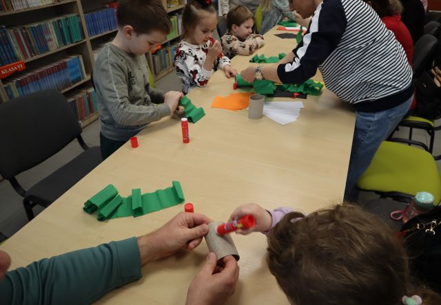 Na zdjęciu znajdują się złączone stoły przy których siedzą dzieci wraz z rodzicami i ozdabiają bibułą rolki po papierze. W tle znajdują się stoły.