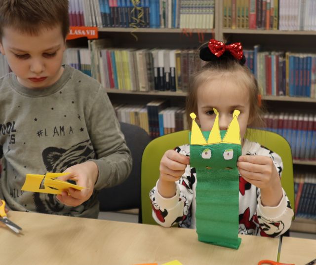 Na zdjęciu znajduje się dziewczynka siedząca przy stoliku i trzyma przed sobą żabę zrobioną z rolki po papierze, ma ona również koronę, po lewej stronie stoi chłopiec i wycina koronę z żółtej kartki. W tle znajdują się półki z książkami.