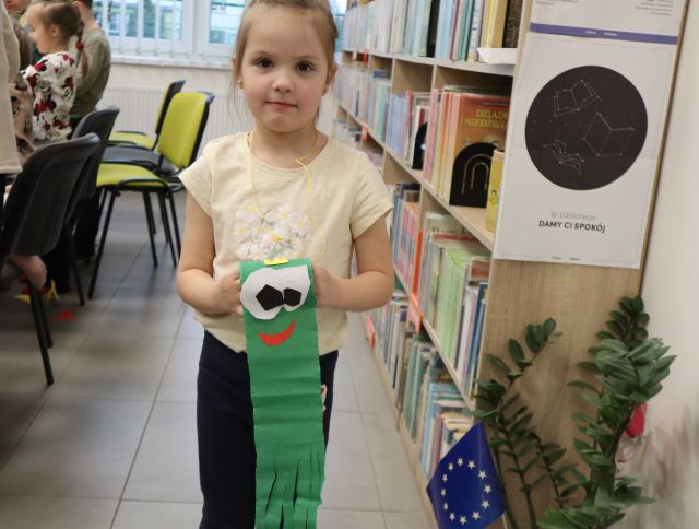 Na zdjęciu znajduje się dziewczynka stojąca między stolikami, a półkami z książkami i trzyma w ręce wykonaną z rolki po papierze i bibuły żabę.