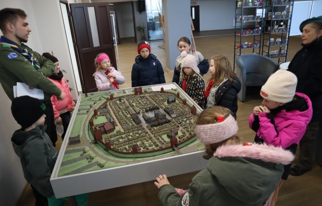 Na zdjęciu znajduje się grupka dzieci oraz dwóch harcerzy i stoją dookoła makiety miasta, która znajduje się na holu biblioteki.