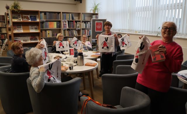 Na zdjęciu znajdują się kobiety na fotelach przy stolikach, dwie z nich stoją i wszystkie trzymają w ręce upominki w postaci białych torb z nadrukiem. W tle znajdują się półki z książkami oraz okna.