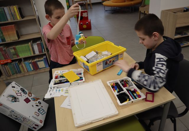 Na zdjęciu znajduje się dwójka chłopców, którzy układają klocki LEGO przy stoliku. W tle znajdują się półki z książkami.