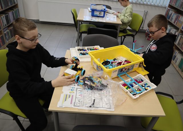 Na zdjęciu znajduje się dwójka chłopców, którzy układają z pudełka klocki LEGO, w tle znajduje się chłopiec, który również układa LEGO. Po lewej i prawej stronie znajdują się półki z książkami.