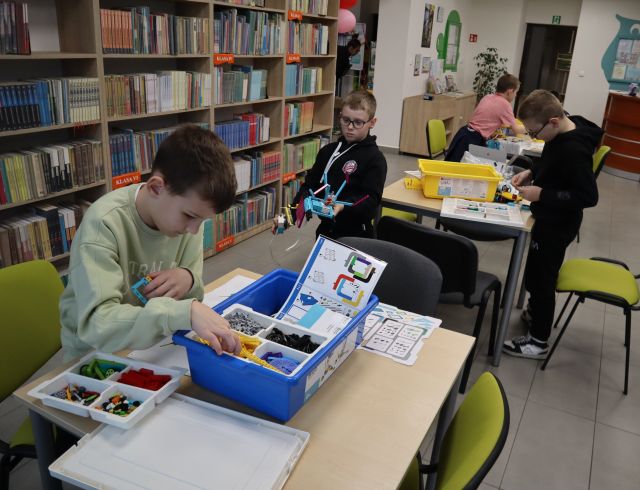 Na zdjęciu znajduje się chłopiec układający LEGO przy stoliku, w tle przy dwóch kolejnych stolikach siedzą chłopcy i również układają LEGO. Po lewej i prawej stronie znajdują się półki z książkami.