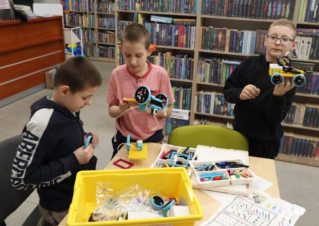 Na zdjęciu znajduje się trzech chłopców, dwóch układa klocki LEGO przy stoliku, a trzeci stoi obok i trzyma w ręce zbudowany przez siebie samochód z LEGO. W tle znajdują się półki z książkami.