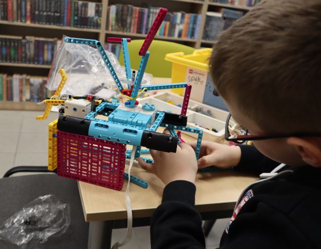 Na zdjęciu znajduje się chłopiec składający konstrukcje z LEGO przy stoliku, na którym leżą klocki i pudełko. W tle znajdują się półki z książkami.