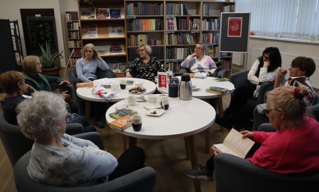 Na zdjęciu znajdują się kobiety siedzące na fotelach przy okrągłych stolikach, na których znajdują się książki, termosy i szklanki. W tle znajdują się półki z książkami.