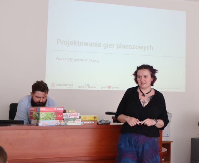 Na zdjęciu znajduje się kobieta, za nią na biurku leżą gry planszowe oraz za biurkiem znajduje się mężczyzna. Na ścianie w tle wyświetlana jest prezentacja.