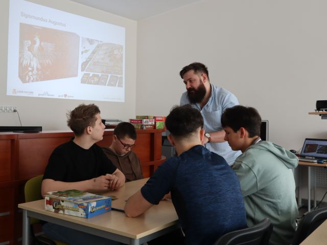 Na zdjęciu znajduje się trójka nastolatków grająca w grę, a obok stolika stoi mężczyzna. Po lewej stronie na ścianie wyświetlana jest prezentacja. Warsztaty odbywają się w sali komputerowej.