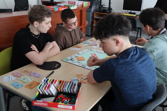 Na zdjęciu znajduje się czterech nastolatków, grających przy stoliku w monopoly. Warsztaty odbywają się w sali komputerowej.
