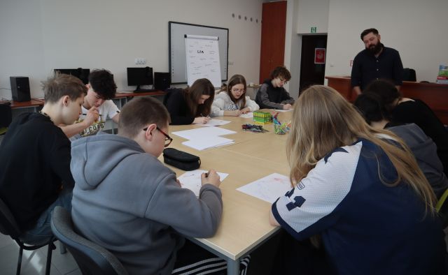 Na zdjęciu znajduje się młodzież przy załączonych stolikach i rysująca na kartkach za pomocą znajdujących się na środku kredek. Obok stolika w tle znajduje się mężczyzna. Warsztaty odbywają się w sali komputerowej.