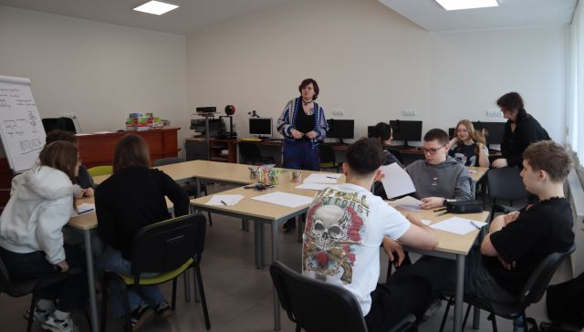 Na zdjęciu znajduje się młodzież przy trzech stolikach, przy których tworzą prototyp swojej gry, na środku znajdują się stoły, na których leżą potrzebne im materiały. W tle znajduje się kobieta. Warsztaty odbywają się w sali komputerowej.