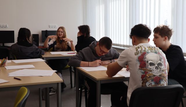 Na zdjęciu w centrum znajduje się trzech chłopaków przy stoliku i tworzą prototyp gry, za nimi znajduje się stolik przy którym siedzą trzy dziewczyny. Pomiędzy nimi po lewej stronie znajdują się stolik z kartkami. Warsztaty odbywają się w sali komputerowej.
