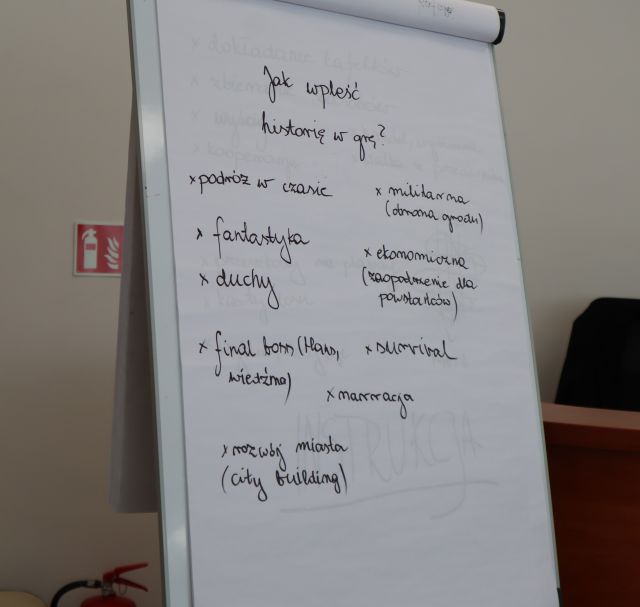 Na zdjęciu znajduje się mobilna tablica znajdująca się w sali, na której wypisane są propozycje jak wpleść historię w grę, na przykład poprzez podróż w czasie, fantastykę, duchy, finał boss, rozwój miasta, narrację, survival, militaria i ekonomię.