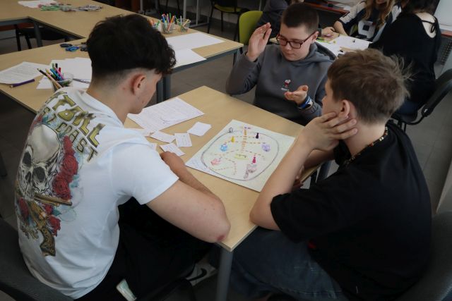 Na zdjęciu znajduje się trójka chłopaków grająca w stworzoną przez nich grę, na kartkach papieru, po lewej stronie znajdują się stoły, na których leżą kartki oraz kredki.